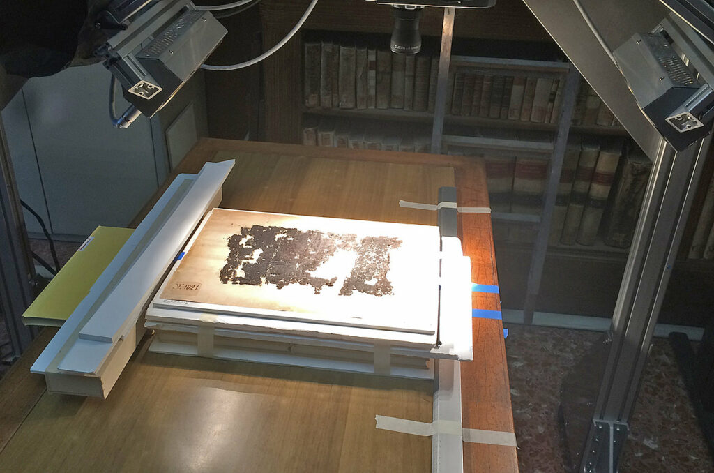Die Fotografie zeigt die Vorbereitungen, um Hyperspektralbilder zu erstellen und die Literatur somit wieder lesbar zu machen. Die Papyrusrolle wird auf einem erhöhten Untergrund platziert und mit Hilfe von Scheinwerfern ausgeleuchtet.