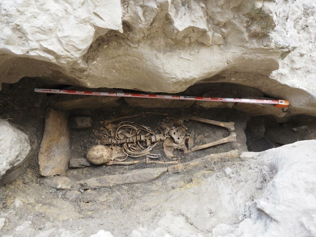Das Foto zeigt das Grab in Ojo Guareña. Zu sehen ist das Skelett in Rückenlage. Das Grab ist mit großen Steinplatten eingefasst. Der Kopf befindet sich neben der Wirbelsäule auf Höhe der Schulter. Die Unterschenkelknochen und Füße sind nicht mehr erhalten.