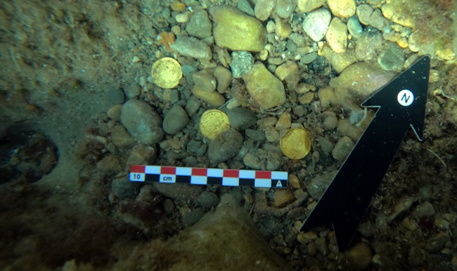Unterwasserfoto von drei Goldmünzen vor der Küste von Portitxol. Alle drei Münzen liegen noch auf dem Meeresboden und sind zum Teil von Steinen leicht bedeckt. Ein Maßstab und ein Nordpfeil wurden für die Fotodokumentation daneben gelegt.
