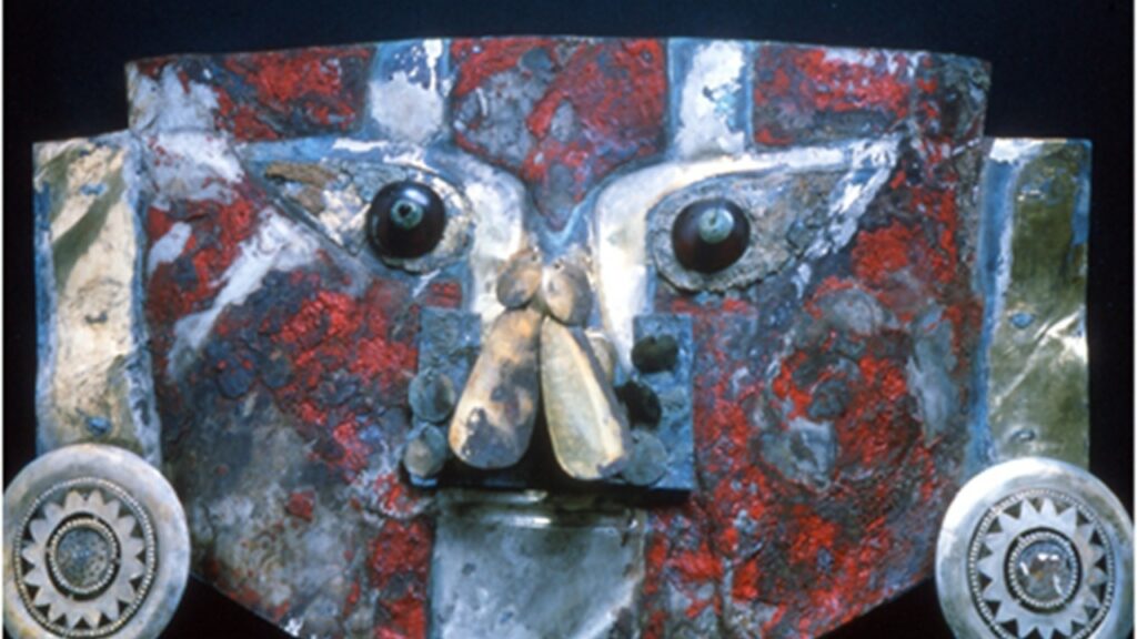Die zum Teil mit roter Farbe bemalte Maske aus dem Grab in Peru zeigt anthropomorphe Gesichtszüge. Auffällig sind die großen runden Scheiben, die Ohrringe darstellen sollen.