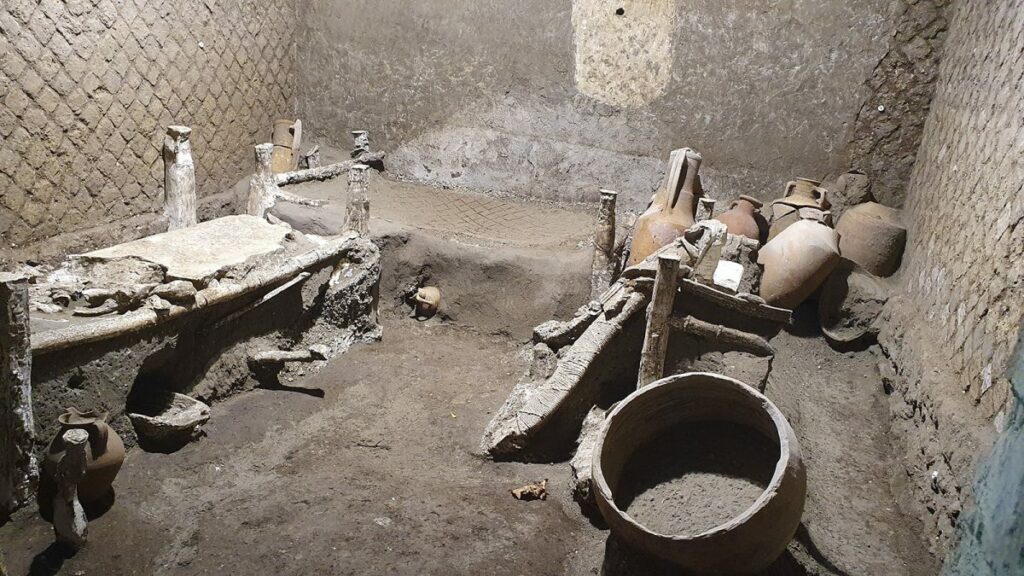Das Bild zeigt den kleinen Raum, in dem die Sklaven der Villa untergebracht waren. Im Raum stehen drei Betten, deren Gestelle noch erhalten sind. In einer Ecke stehen große Amphoren.
