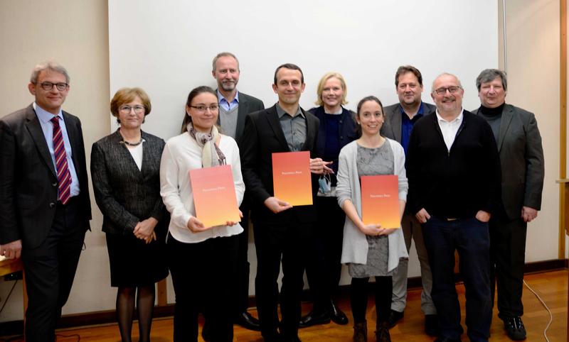 Die drei Nachwuchswissenschaftler, die mit dem Philippika-Preis ausgezeichnet wurden, stehen in der Mitte und halten einen Umschlag in orange und roten Farbtönen. Hinter ihnen stehen die restlichen Beteiligten der Preisverleihung.