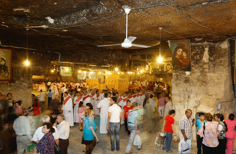 Grotte in der Nähe des Klosters Deir al-Adra, in der die Heilige Familie während ihrer Flucht stationierte. Die Aufnahme zeigt einen mit Glühbirnen ausgeleuchteten großen, steinernen Raum, in dem sich viele Menschen befinden. An den Wänden hängen Bilder der Heiligen Maria und dem Jesuskind sowie Bilder der Heiligen Familie.