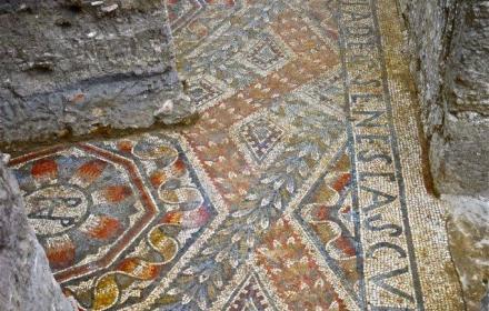 Detail des Mosaiks aus der Villa Romana. Zu sehen ist ein Teil des Randes mit einer Inschrift sowie geometrischen Formen.
