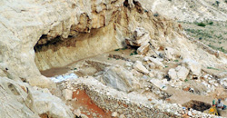 Zu sehen ist ein Felsüberhang im Jebel Faya unter und vor dem Ausgrabungen statt finden. Deutlich sind einzelne Grabungsschnitte und Grabungsarbeiter zu erkennen.