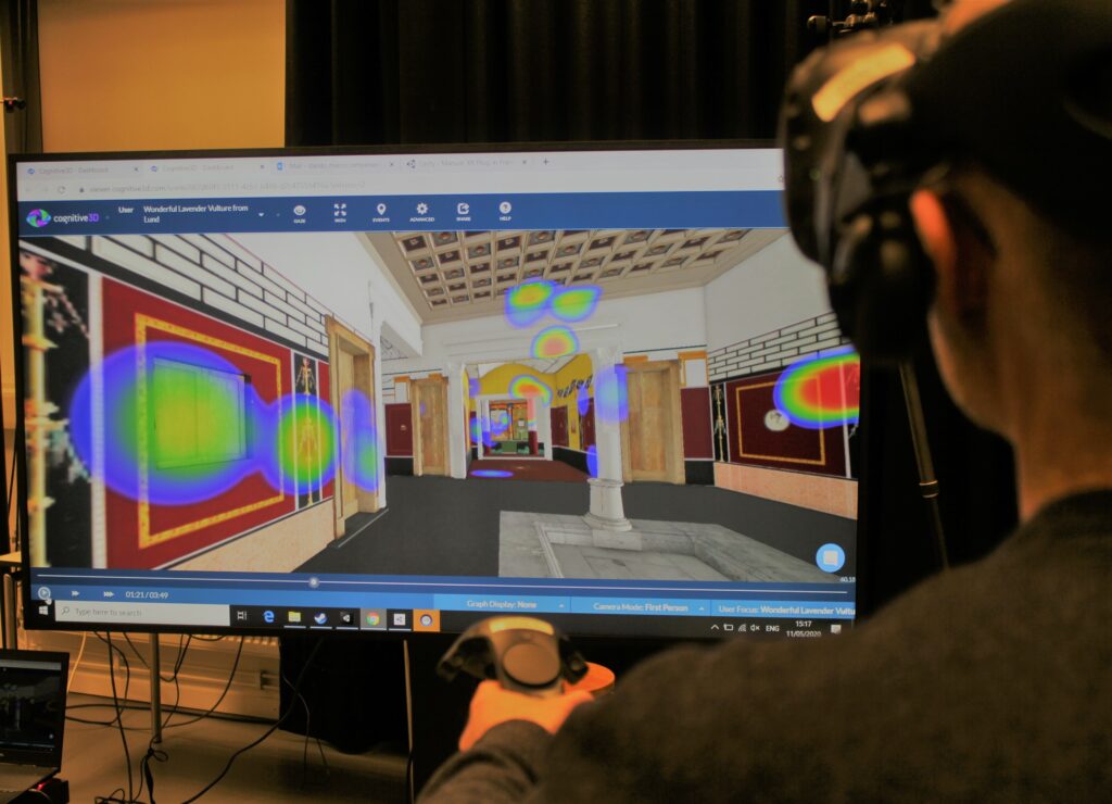 Das Bild zeigt eine Probandin, die mit Hilfe einer VR-Brille und einem Joystick durch das römische Haus geht. Vor ihr befindet sich ein Bildschirm, der das Haus in Virtual Reality zeigt. Die Bereiche, die näher angesehen werden, sind farblich hervorgehoben (z.B. Wandmalereien oder Decken).