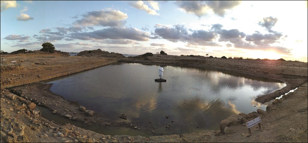 Panoramaaufnahme des Beckens in Mozia, das mit Wasser aufgefüllt wurde. In der Mitte befindet sich die Replik einer Ba'al-Statue. Zu sehen ist der Torso sowie ein Arm.