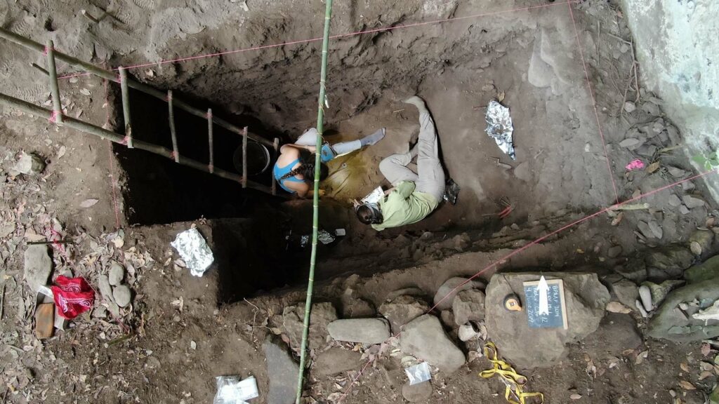 Blick von oben in einen Grabungsschnitt unterhalb eines Felsüberhangs. Zwei Archäologen sitzen auf dem Boden und legen die das Skelett eines Einwanderers frei, der womöglich Mais nach Mittelamerika brachte.