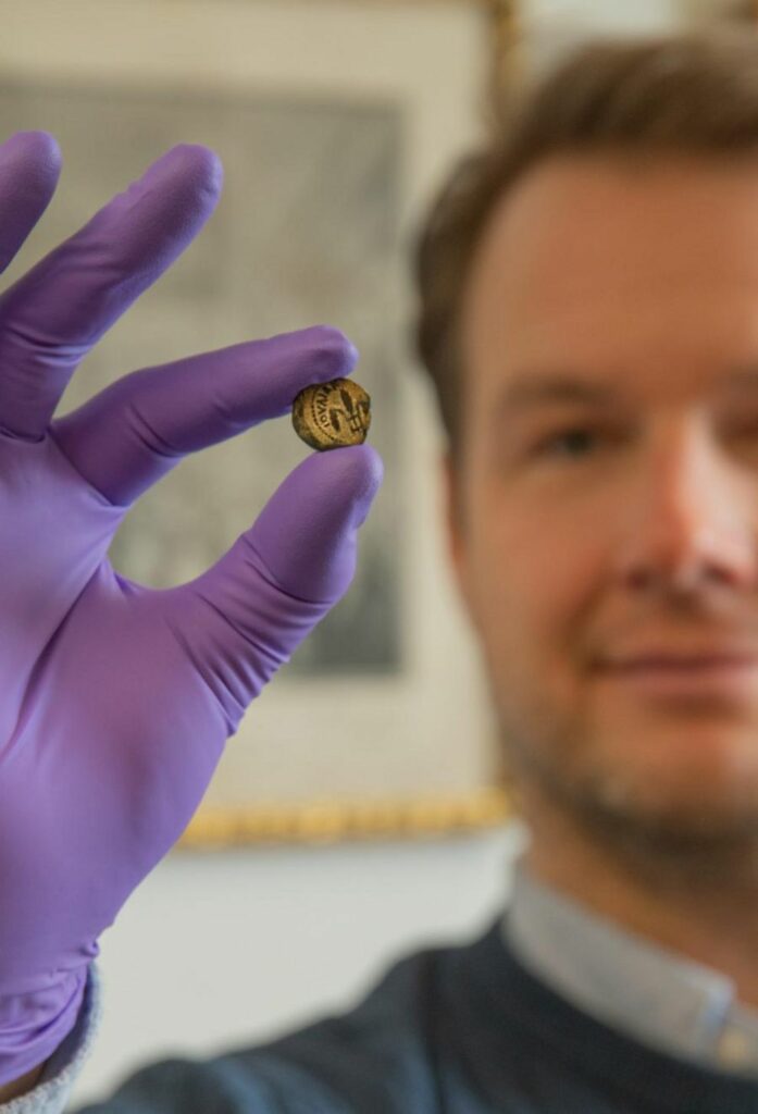 Professor Stefan Krmnicek hält eine der 394 Münzen in seiner behandschuhten Hand. Die kleine Münze (ca. Größe 1 Cent) ist von gelblicher Farbe. Sie zeigt eine stilisierte Blume, die an eine Lilie erinnert.