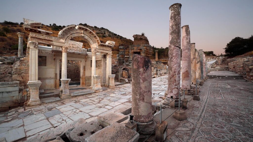 Das Foto zeigt die Front des Hadrianstempel an einer Straße in Ephesos. Erhalten sind noch der Arkadenbogen und das Eingangsportal, sowie die Platten der Straße. Gegenüber befand sich ein Gebäude mit einer Säulenstellung als Fassade und einem Mosaikboden.