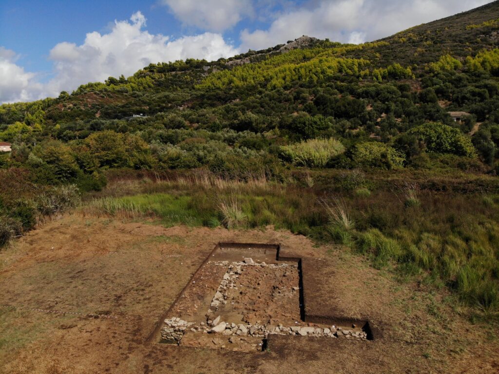 Blick in die Landschaft von Elis: Grabungsschnitt mit Fundamentresten im Vordergrund, die Akropolis von Samikon im Hintergrund