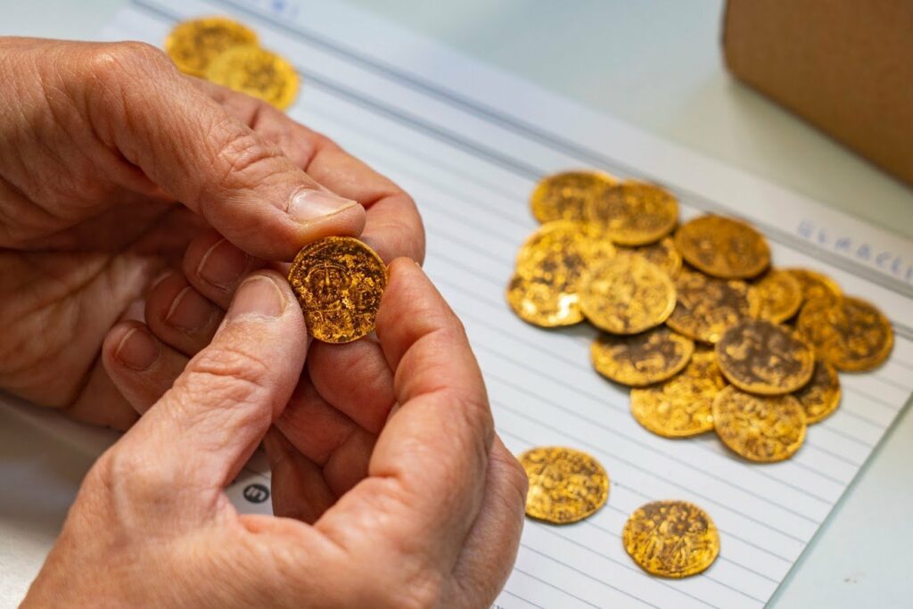 Byzantinische Goldmünzen liegen auf einem Haufen. Im Vordergrund hält ein Paar Hände eine Goldmünze