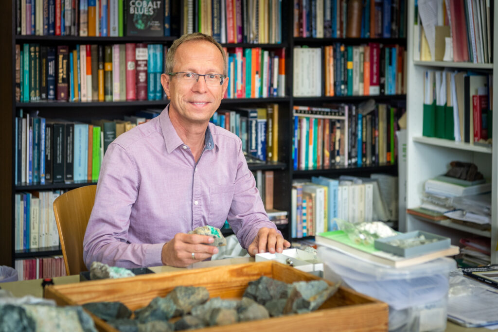 Prof. Albert Gilg sitzt vor einem Bücherregal an einem Schreibtisch und hält einen Stein in der Hand