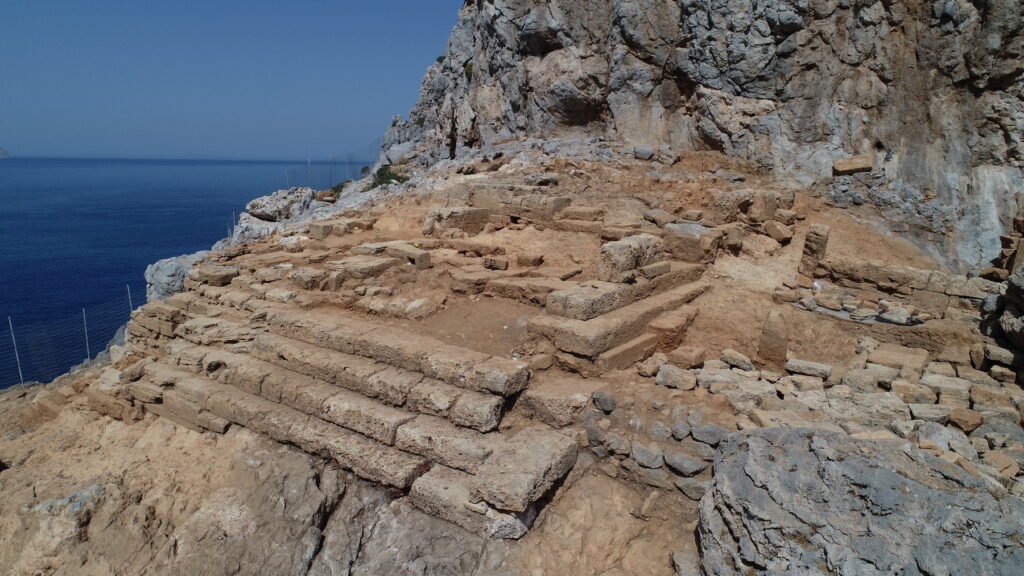 Ansicht des Tempels im Demeter-Heiligtum von Falassarna, Kreta. Der Tempel, von dem das Fundament mit einer großen Freitreppe erhalten ist, steht vor einer steilen Felswand. Im Hintergrund ist das Meer zu sehen.