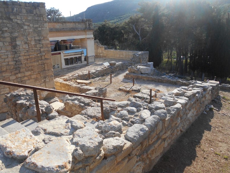Foto des Wegs zu den Megara des Königs und der Königin im Palast von Knossos. Hinter einer alten Mauer aus großen Steinen sieht man eine Treppe und einen verwinkelten Weg, der an einer Seite abgezäunt ist.