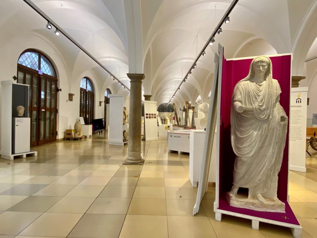 Blick in das Römische Museum Augsburg. Zu sehen ist die Wanderausstellung "Antike in Bayern. Im Vordergrund steht eine Statue des römischen Kaisers Augustus in einer mit rotem Stoff ausgekleideten Kiste.
