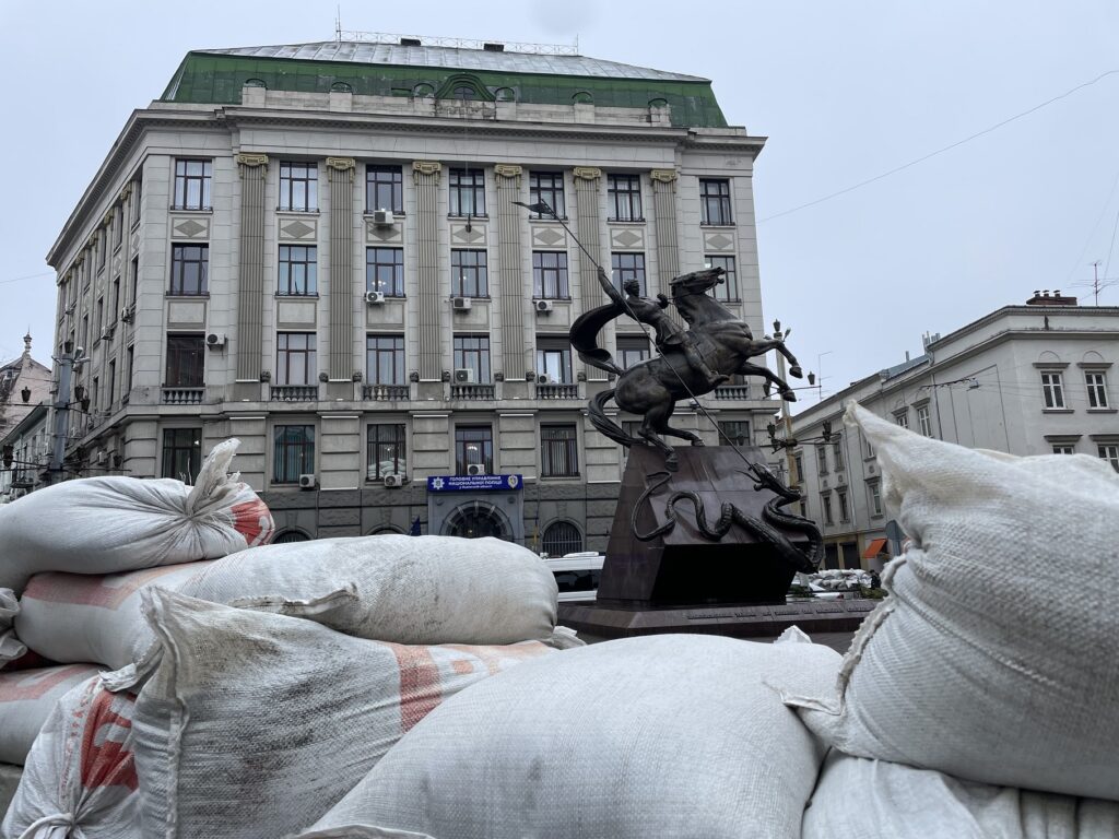 Schutzmaßnahmen an einem Momument in Lviv. Im Vordergrund liegen Sandsäcke, dahinter sieht man eine bronzene Statue eines Reiters, der gegen eine Schlange kämpft, und ein klassizistisches Gebäude.