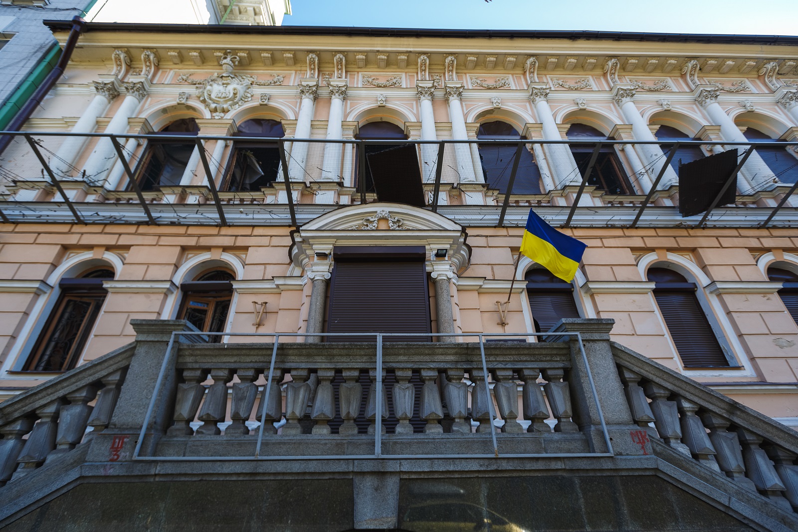Eingang des Khanenko Museums in Kiew: neben dem geschlossenen Eingang hängt die ukrainische Flagge, einige Fenster sind geöffnet, andere mit Rollläden verschlossen. Zwischen den beiden Etagen ist ein Netz gespannt, auf dem zwei große dunkle Bretter liegen, die ursprünglich die Fenster verschlossen hatten.