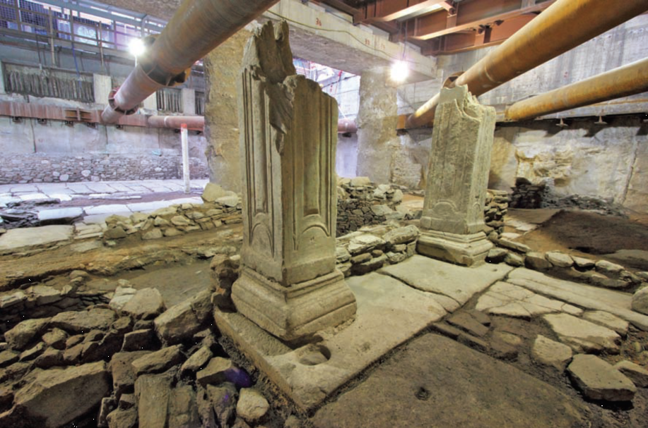 Foto von der Ausgrabung im Venizelos-Bahnhof Thessaloniki: zu sehen ist eine Türschwelle, die von den Resten zweier Pfeiler flankiert ist. Oberhalb der antiken Reste verlaufen moderne Rohre.